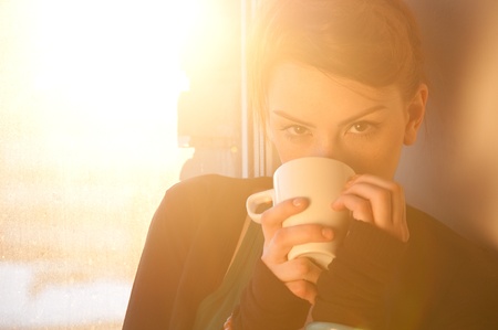 Beautiful young woman in casual wear dreaming with cup of hot coffee over window. Sun beam.の写真素材