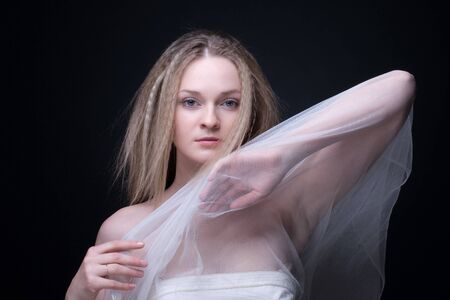 Closeup portrait of beautiful blond girl with blue eyes in white fabric on blackの写真素材
