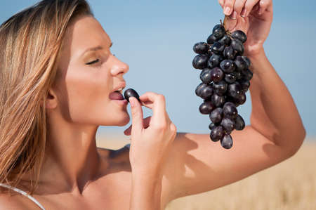 Beautiful woman with perfect hair and skin posing in wheat field and eating grapes. Summer picnic.の写真素材