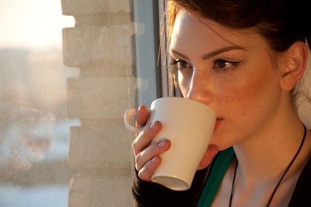 Beautiful young woman in casual wear dreaming with cup of hot coffee over window