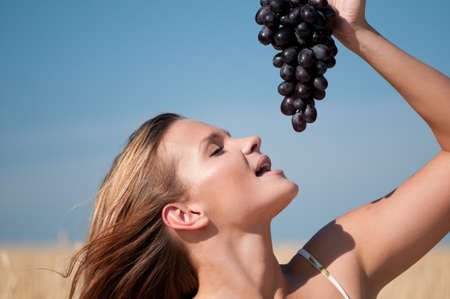 Beautiful woman with perfect hair and skin posing in wheat field and eating grapes. Summer picnic.の写真素材