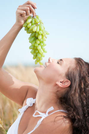 Beautiful woman with perfect hair and skin posing in wheat field and eating green grapes. Picnic.の写真素材