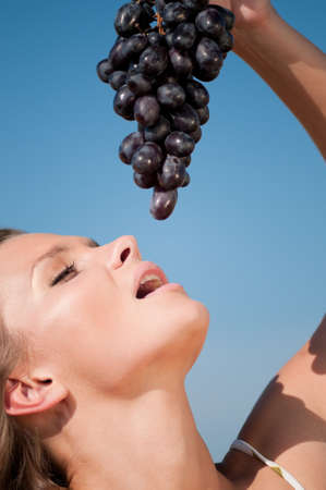 Beautiful woman with perfect hair and skin posing in wheat field and eating grapes. Summer picnic.の写真素材