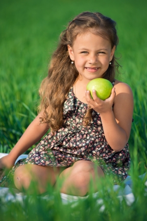 Happy girl with apple on green grass at spring or summer park picnicの写真素材