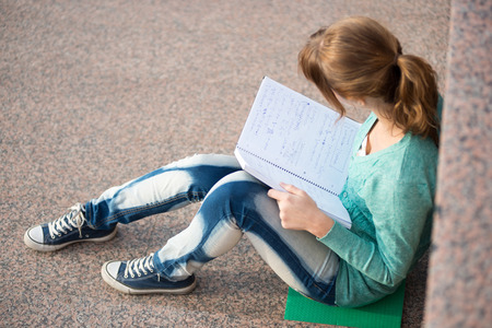 Portrait of a beautiful teenage student girl sitting on stairs in park with copy book and studing. Sunny summer day. Modern and casual lifestyle. Woman with note pad.の写真素材