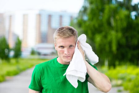 Tired man with white towel after fitness time and exercising in city street park at beautiful summer day. Sporty model caucasian ethnicity training outdoor.の写真素材