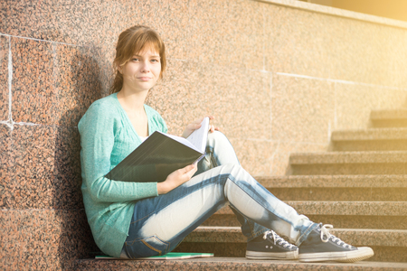 Portrait of a beautiful teenage student girl sitting on stairs in park with copy book and studing. Sunny summer day. Modern and casual lifestyle. Woman with note pad.の写真素材
