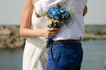 the bride hugs the groom and holds a Bridal bouquet with blue flowers on a background of the riverの写真素材