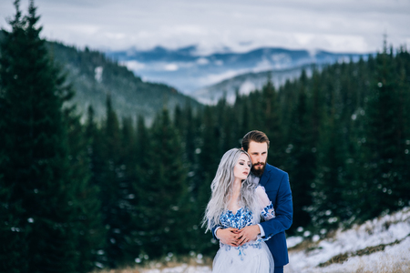 groom in a blue suit and bride in white, embroidered with blue pattern, dress on a background of green pine forests in the mountains of the Carpathiansの写真素材
