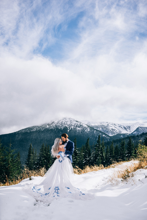groom in a blue suit and bride in white, embroidered with blue pattern, dress on a background of green pine forests in the mountains of the Carpathiansの写真素材