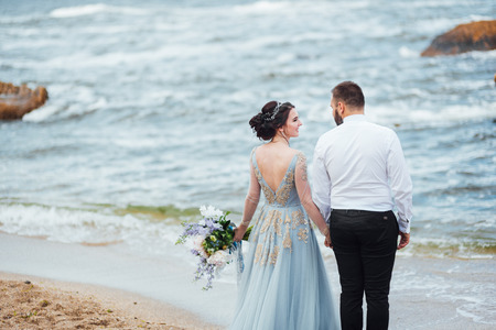 same couple with a bride in a blue dress walk along the ocean shoreの写真素材