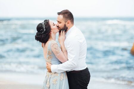 same couple with a bride in a blue dress walk along the ocean shoreの写真素材