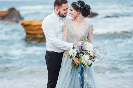 same couple with a bride in a blue dress walk along the ocean shoreの写真素材