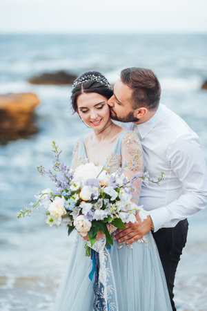 same couple with a bride in a blue dress walk along the ocean shoreの写真素材