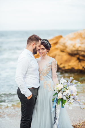 same couple with a bride in a blue dress walk along the ocean shoreの写真素材