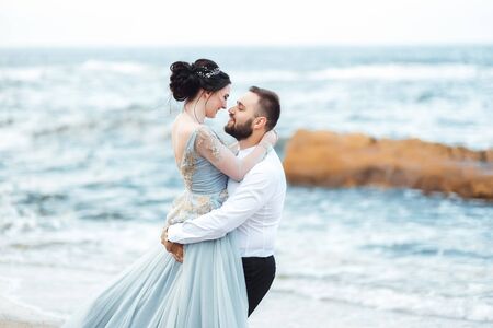 same couple with a bride in a blue dress walk along the ocean shoreの写真素材