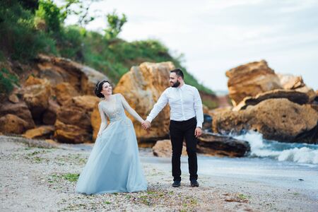 same couple with a bride in a blue dress walk along the ocean shoreの写真素材