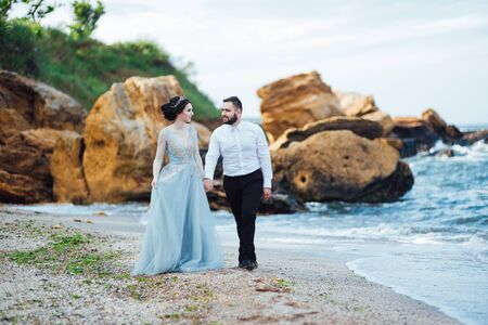 same couple with a bride in a blue dress walk along the ocean shoreの写真素材