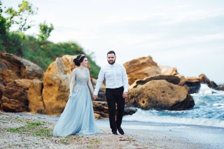 same couple with a bride in a blue dress walk along the ocean shoreの写真素材