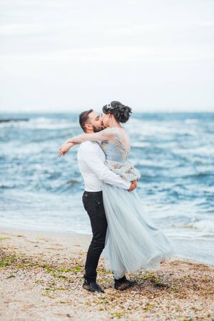 same couple with a bride in a blue dress walk along the ocean shoreの写真素材