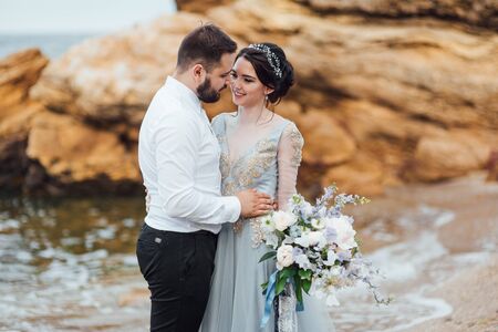 same couple with a bride in a blue dress walk along the ocean shoreの写真素材