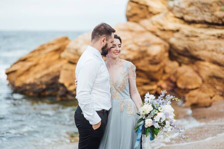 same couple with a bride in a blue dress walk along the ocean shoreの写真素材