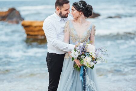same couple with a bride in a blue dress walk along the ocean shoreの写真素材
