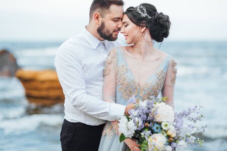 same couple with a bride in a blue dress walk along the ocean shoreの写真素材