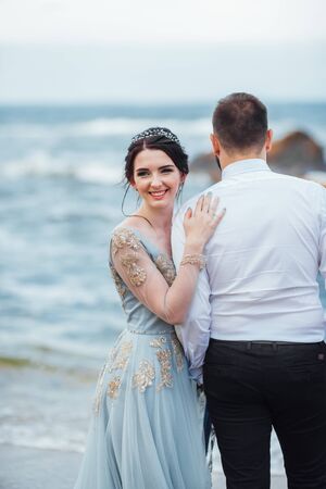 same couple with a bride in a blue dress walk along the ocean shoreの写真素材
