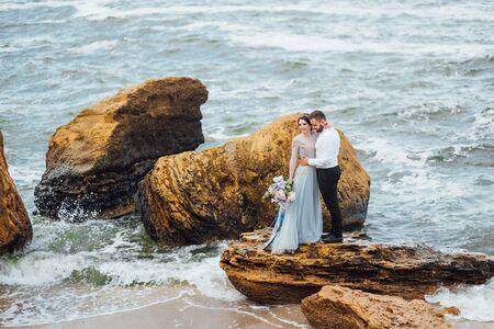 same couple with a bride in a blue dress walk along the ocean shoreの写真素材