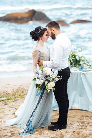same couple with a bride in a blue dress walk along the ocean shoreの写真素材