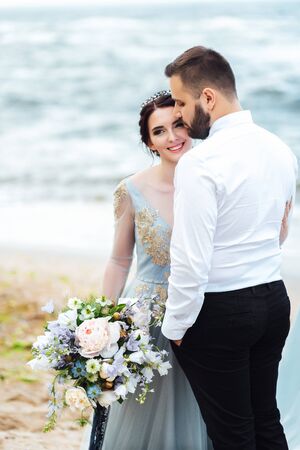 same couple with a bride in a blue dress walk along the ocean shoreの写真素材