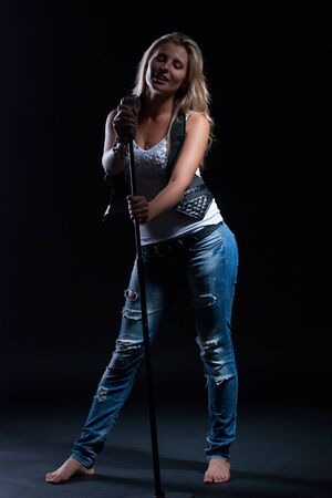 young, girl, blonde, jeans, clothes on a dark background emotionally sings into a microphoneの写真素材