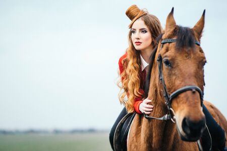 red-haired jockey girl in a red cardigan and black high boots with a horse for a walkの写真素材