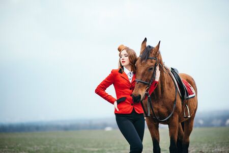 red-haired jockey girl in a red cardigan and black high boots with a horse for a walkの写真素材