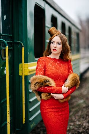 young girl with red hair in a bright red dress near an old green passenger carの写真素材