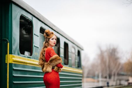 young girl with red hair in a bright red dress near an old green passenger carの写真素材