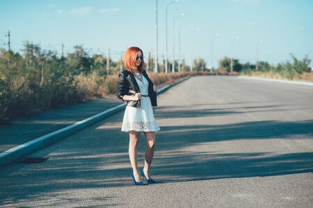 red-haired girl in a black jacket and blue glasses on a gray roadの写真素材