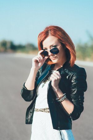 red-haired girl in a black jacket and blue glasses on a gray roadの写真素材