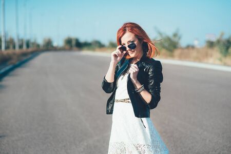 red-haired girl in a black jacket and blue glasses on a gray roadの写真素材