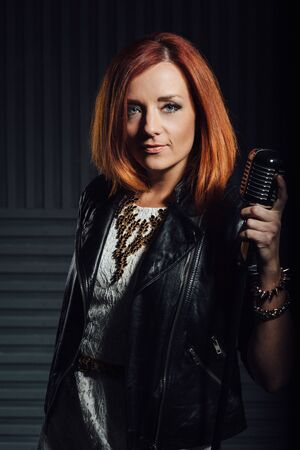 young red-haired girl in a white dress and a black leather rocker jacket sings emotionally into the microphoneの写真素材