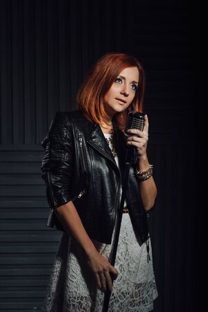 young red-haired girl in a white dress and a black leather rocker jacket sings emotionally into the microphoneの写真素材