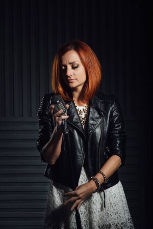 young red-haired girl in a white dress and a black leather rocker jacket sings emotionally into the microphoneの写真素材