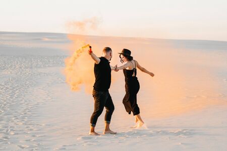 guy and a girl in black clothes hug and run on the white sand with orange smoke