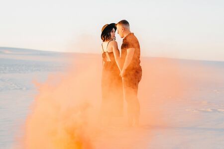 guy and a girl in black clothes hug and run on the white sand with orange smokeの写真素材