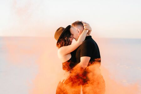 guy and a girl in black clothes hug and run on the white sand with orange smokeの写真素材