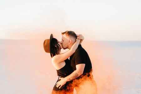 guy and a girl in black clothes hug and run on the white sand with orange smokeの写真素材