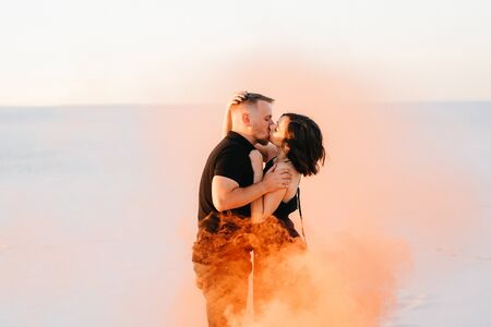 guy and a girl in black clothes hug and run on the white sand with orange smokeの写真素材