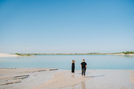 young couple a guy with a girl in black clothes are walking on the white sand at the edge of blue waterの写真素材