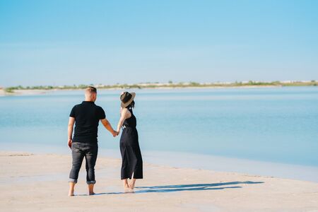 young couple a guy with a girl in black clothes are walking on the white sand at the edge of blue waterの写真素材